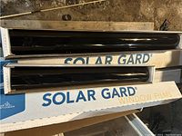 Black tinted window film rolls in Solar Gard labeled boxes, one roll open to show film color and texture.
