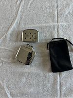 Overhead photo showing Zippo hand warmer opened with vented cover removed and black pouch nearby on white fabric background.