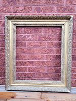 Full front view of the large antique picture frame with ornate gold finish against a brick wall.