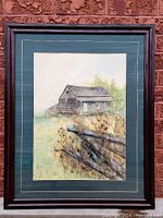 Framed watercolour painting depicting a rustic barn and fence in natural surroundings with matting and wooden frame.