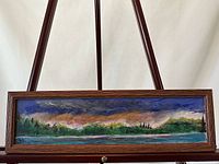 Framed landscape oil painting on board depicted horizontally on an easel. The wood frame is visible with the image showing a river, trees and a colorful cloudy sky.