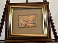 Framed sterling silver embossed art showing a cottage scene inside gold-toned wooden frame with beige matte