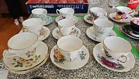 Photo showing most of the teacup and saucer pairs arranged on a table, various floral patterns and styles visible.