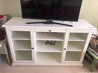 Front view of the white wooden cabinet with a TV on top. Cabinet has a central section with open shelves and two side sections with glass doors.