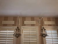 Overall view of three rectangular wooden wall signs reading 'dream', 'believe', and 'enjoy' mounted above windows with shutters and two hanging candle holders below them.