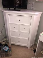 Front view of white corner dresser with five drawers and metal knobs, showing the beveled top and angled shape.