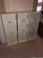 Photo showing the front view of three white manufactured file organizers with four drawers each, placed side by side on a carpeted floor near a wall.