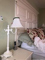 Two white matching table lamps against a wall near a bed. One lamp is fully visible with fabric lampshade and ornate metal body and base.