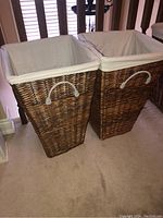 Two wicker laundry hampers with white cotton liners standing side by side on carpeted floor near stair railing. Each hamper has white handles. Photos taken to show condition and details of the liners and handles.