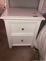 Front view of white nightstand showing two drawers with round metal knobs, placed against wall on carpet.