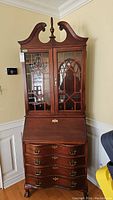 Front view of tall wooden secretary cabinet with bonnet top and glass paneled doors above fall front desk with curved drawers below