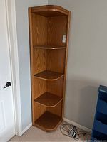 Full view of the tall wooden corner shelf unit showing its size and shape placed in a room corner next to a door and a blue furniture piece.