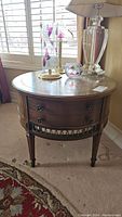 Front view of the vintage wood round side table showing one drawer, metal ring pulls, decorative metal accents, and brass capped feet with wear on top finish.