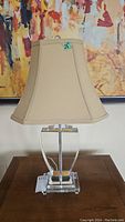 Front view of one table lamp showing clear glass base with square pedestal and beige fabric shade with stitched seams.