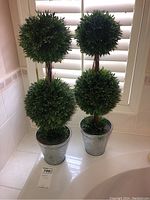 Two faux plant topiaries displayed side by side on a white surface near window blinds, showing overall height and pots.