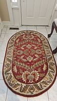 Top view of oval wool rug with red, beige, and brown floral pattern in entryway on white tiled floor.