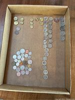 Top-down view showing many foreign coins arranged in rows inside a cardboard box, including euro coins of different denominations and colors.