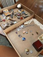 Overview of two cardboard trays with assorted costume jewelry and accessories, including brooches, pendants, rosaries, a pen, a clock, and small containers.