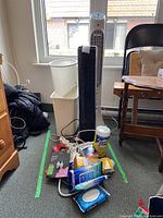 Photo showing white Honeywell tower fan and black Sunbeam tower fan, two white waste baskets, various cleaning supplies and power strips on floor.