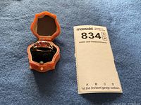 Photo showing 14K yellow gold ring with 3 garnets set horizontally, placed inside an orange vintage-style octagonal jewelry box with black velvet interior.