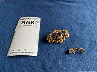 Gold filled pin with three round faceted amethyst stones and ornate gold scrollwork beside a pair of 14K gold and white metal shell shaped clip-on earrings lying flat on a blue fabric.