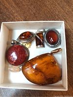 All six jewelry pieces arranged in a small white box. Pieces include amber pendants and brooch, polished stones, and silver settings.