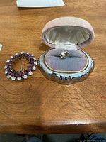 Sterling silver circular pin with purple stones and clear rhinestones next to 10K gold ring in pink velvet-lined box.