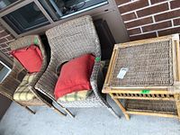 Two faux wicker chairs with red and plaid seat cushions and one faux wicker side table, all shown on patio floor beside brick wall.