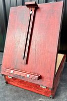 Front view of the wooden art easel showing adjustable vertical support, bottom ledge, and red distressed finish.