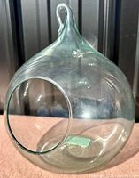 Clear blown glass terrarium with a rounded shape and hook for hanging, shown from the front with the opening visible.