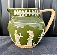 Front view showing three Grecian figures, an urn on pedestal, and leaf vine border at top on olive green glossy ceramic pitcher's body and cream-colored handle.