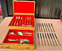 Wide shot showing two-level wooden chest with red felt lining containing gold plated flatware pieces and additional knives laid out.