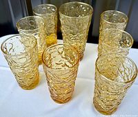 Nine vintage amber/golden crinkle texture pressed glass tumblers arranged in a group on white cloth showing textured pattern and variety in height.