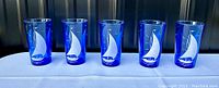 Five cobalt blue glass tumblers with white etched sailboat designs displayed side by side on a light blue fabric surface.