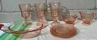 Overview photo showing all pink depression glass items including stemmed goblets, bowls, and plates arranged on a white and green striped cloth.