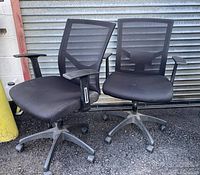 Two black rolling office chairs with mesh backs, padded fabric seats, and armrests shown from the front, placed outside on asphalt near a metal door and yellow pole.