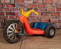Front-left angled view of the Big Wheels tricycle showing large black rubber front wheel with silver spokes, yellow handlebar, blue seat, and orange frame.