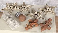 Photo showing four beige barn stars, rusty metal ball, small metal scallop shell plaque, rectangular anchor plate with urn design, and two rusted fleur-de-lis finials arranged on a table.