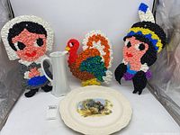 Front view of all items: pilgrim woman, Native American woman, turkey popcorn plastic decorations, ceramic turkey platter, and white ceramic pitcher vase.