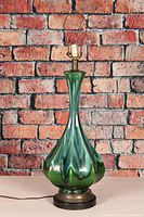 Full view of mid century ceramic lamp with green drip glaze body and brass-trimmed base against brick wall background.
