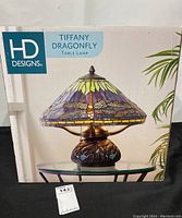 Box showing front of HD Designs Tiffany Dragonfly Table Lamp with depiction of stained glass style lamp and dragonfly design.