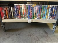 Photo showing a collection of VHS tapes and DVDs on the lower shelf of a metal cart. Titles from animated and family movies are visible, arranged side-by-side.