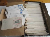 Open box filled with stacked first day cover envelopes and a few laid-out examples showing assorted stamps