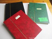 Photo showing three hardcover stamp albums in red, black, and green covers placed on a wooden surface.