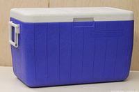 Front and side view of blue rectangular Coleman cooler with white lid and handle and closeable drain on the side.