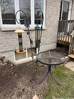 Two metal bird feeders on black hooked stands and a small round metal mesh patio table outside near a brick wall.