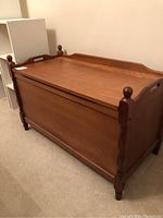 Front angled view of wooden blanket box showing solid wood top with raised edges, plywood sides, rounded corner posts with finials, and turned legs.