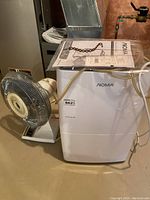 NOMA dehumidifier and Mastercraft table fan positioned side by side on a basement floor showing included owner's manual on top of the dehumidifier.