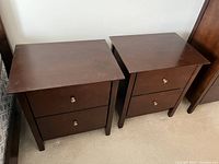 Pair of matching wooden end tables side by side showing tops and fronts with two drawers each and metal knobs, dark brown finish with surface wear.