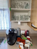 Keurig B60 coffee maker on countertop with assorted mugs and water bottles around it
